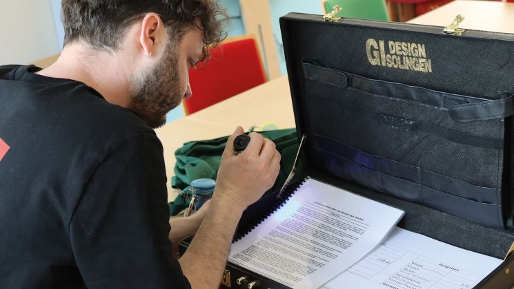 Ein Mann arbeitet konzentriert mit einem Dokument in einer Aktentasche und einer Lupe. Der Hintergrund zeigt bunte Stühle.