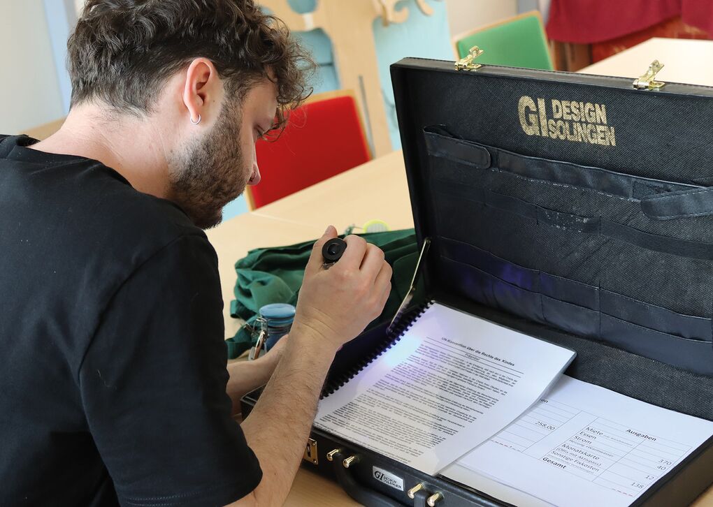 Ein Mann arbeitet konzentriert mit einem Dokument in einer Aktentasche und einer Lupe. Der Hintergrund zeigt bunte Stühle.