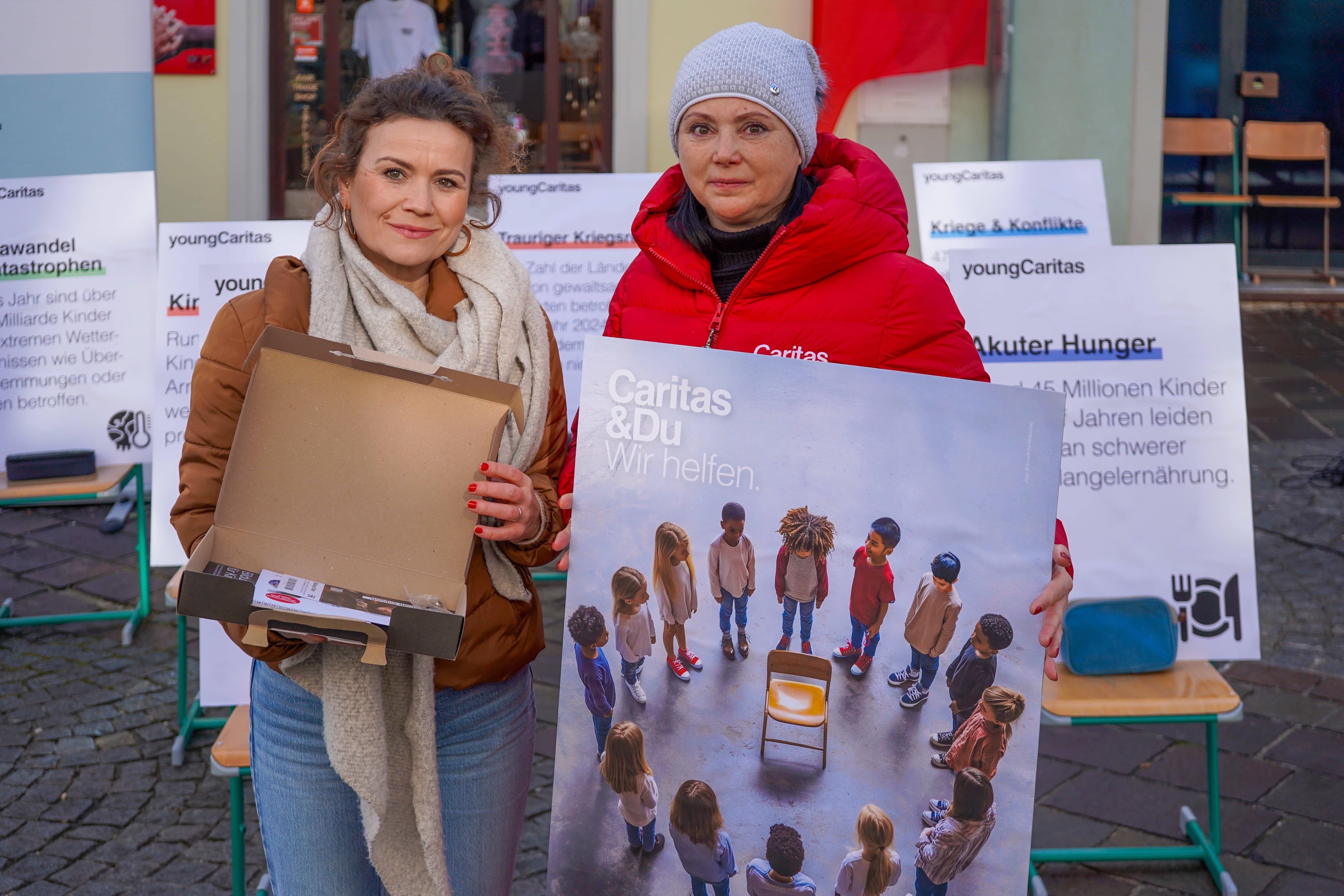 Zwei Frauen präsentieren ein Plakat von Caritas, um auf den Hunger von Kindern aufmerksam zu machen. Hintergrund mit weiteren Ankündigungen.