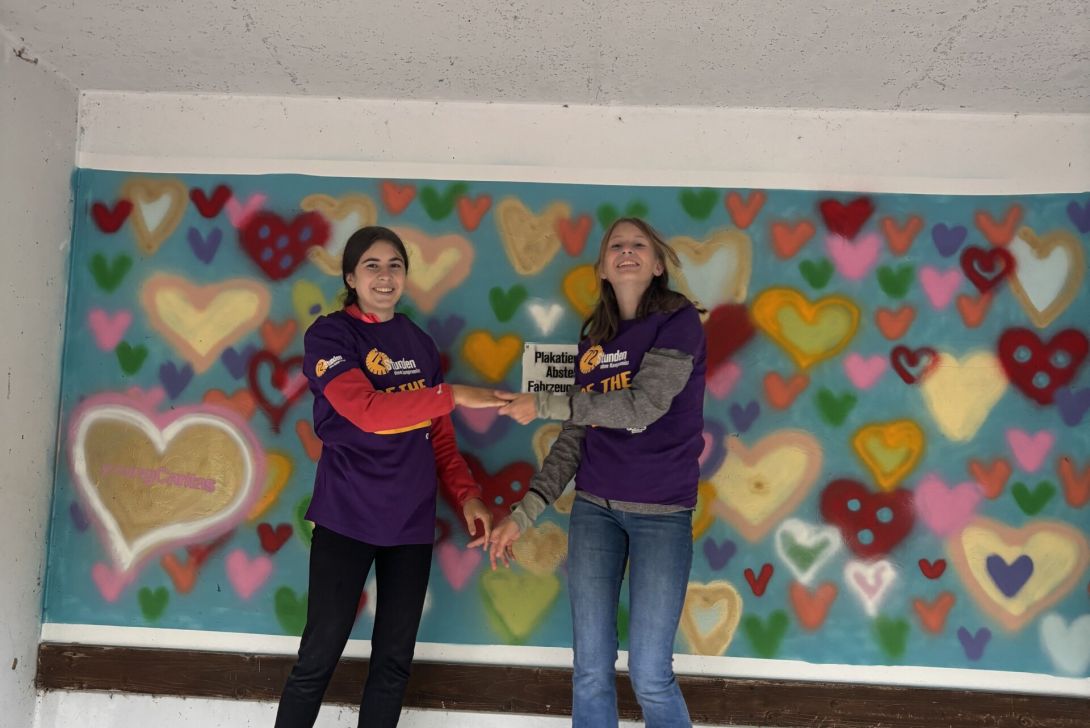 Zwei Frauen stehen in bunten Shirts an einer bunte Herz-Wand. Sie lächeln und haben Spaß in einer kreativen Umgebung.