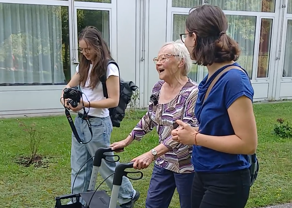 Eine ältere Frau benutzt einen Rollator im Freien, um mit zwei jungen Frauen zu interagieren. Sie lachen und genießen die Zeit.