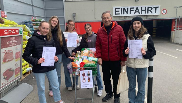Eine Gruppe von jungen Frauen und einem Mann hält Informationsblätter vor einem Supermarkt mit einem Einkaufswagen voller Lebensmittel.