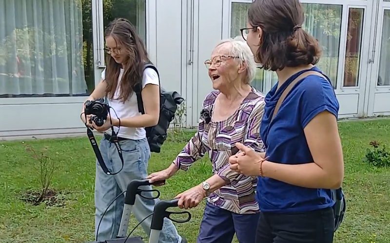 Eine ältere Frau benutzt einen Rollator im Freien, um mit zwei jungen Frauen zu interagieren. Sie lachen und genießen die Zeit.