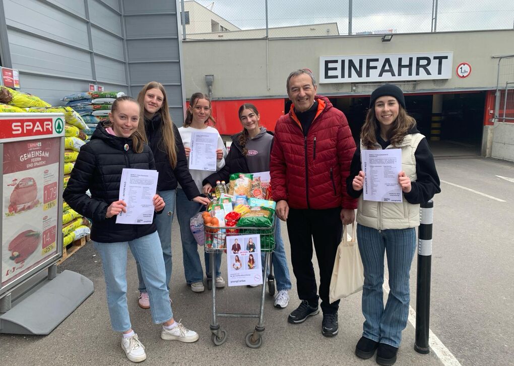 Eine Gruppe von jungen Frauen und einem Mann hält Informationsblätter vor einem Supermarkt mit einem Einkaufswagen voller Lebensmittel.