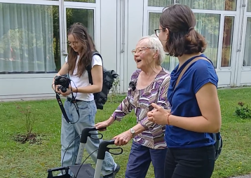 Eine ältere Frau benutzt einen Rollator im Freien, um mit zwei jungen Frauen zu interagieren. Sie lachen und genießen die Zeit.