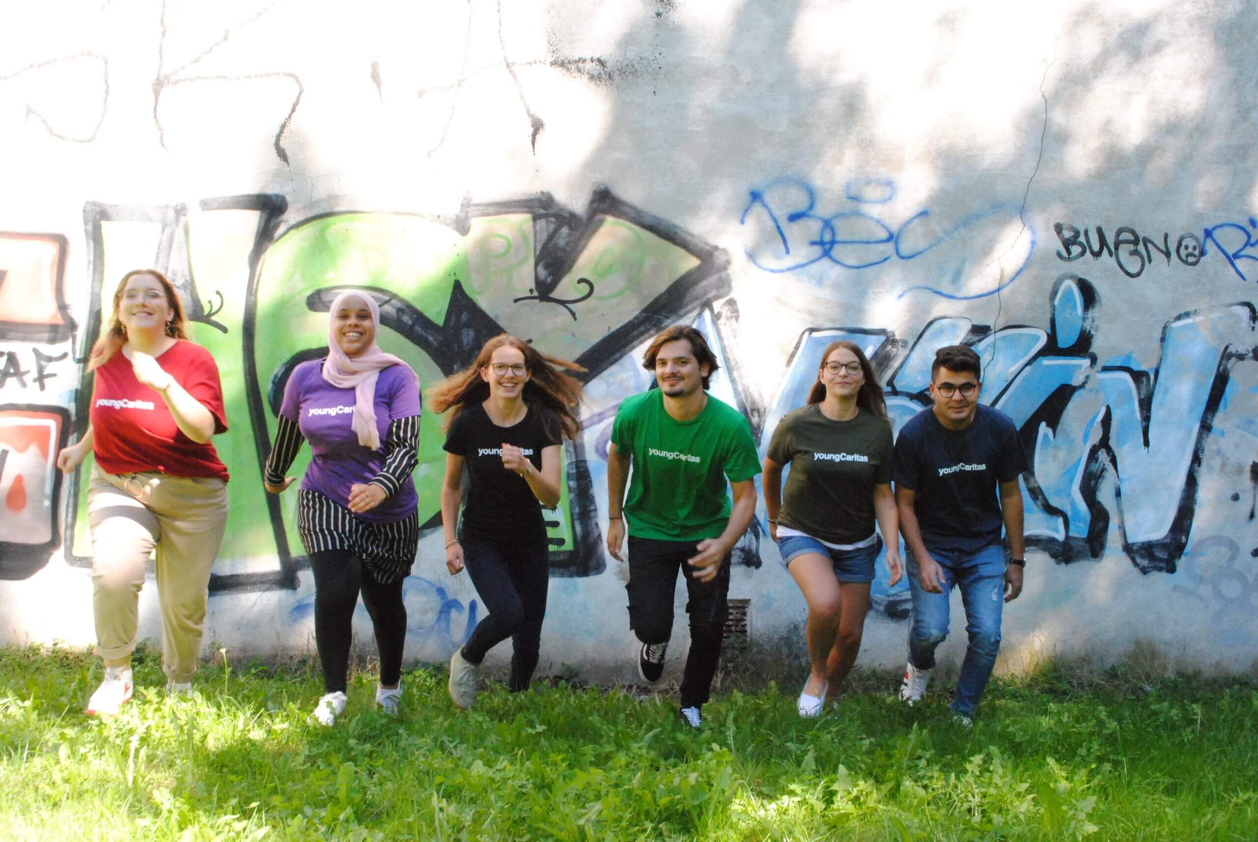 Sechs junge Menschen in bunten T-Shirts rennen fröhlich vor einer Graffiti-Wand im Freien.