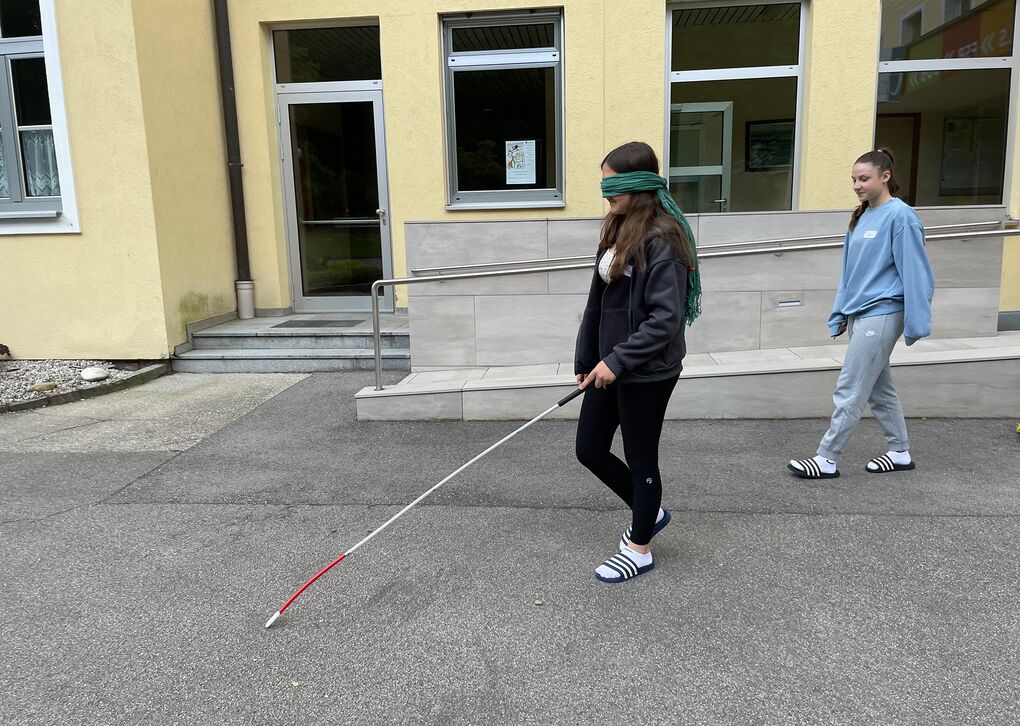 Eine Person mit verbundenen Augen benutzt einen Langstock, während eine andere Person sie begleitet. Sie befinden sich in einem Schulhof.