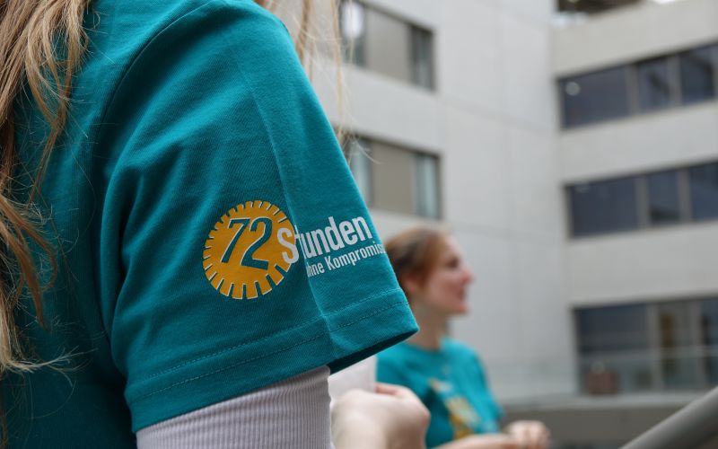 Detailansicht eines T-Shirts mit dem Logo von '72 Stunden ohne Kompromiss', in einer Outdoor-Umgebung mit Menschen im Hintergrund.