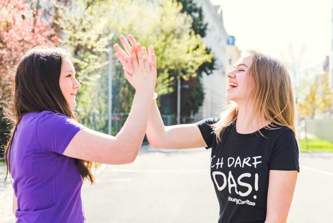 Zwei junge Frauen in sonniger Umgebung geben sich fröhlich High-Fives, während sie lachen.
