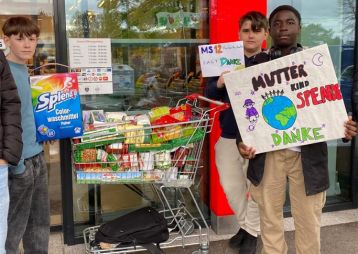 Drei Jugendliche stehen vor einem Supermarkt mit einem Einkaufswagen voller gespendeter Lebensmittel und halten Schilder, auf denen 'Danke' steht.