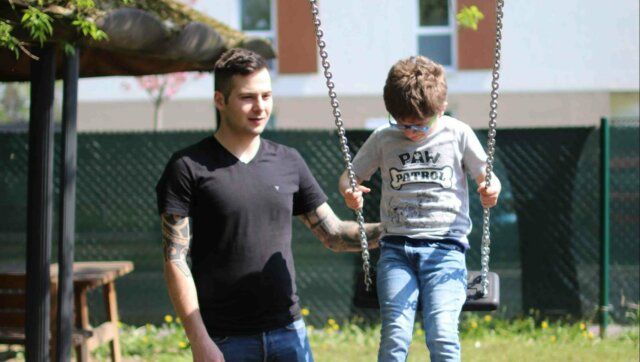 Ein Vater schiebt seinen Sohn auf einer Schaukel im Freien, umgeben von fröhlicher Natur und dem Spielplatz.
