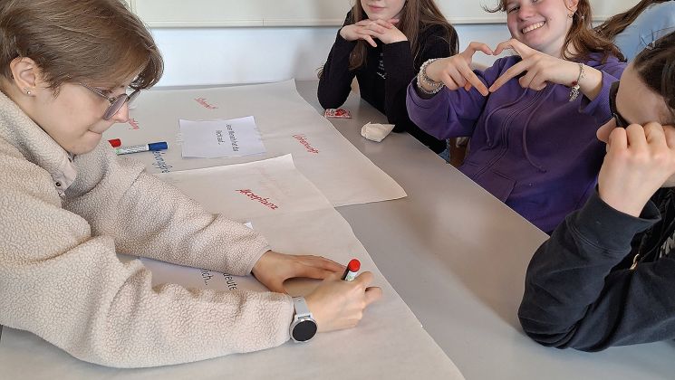 Vier Schülerinnen arbeiten gemeinsam an einem Tisch mit Plakaten und Stiften, während eine von ihnen lächelt und ein Herz formt.