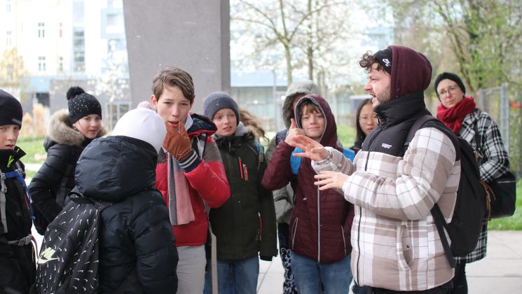 Eine Gruppe von Schülern steht draußen und hört einem Lehrer bei einer Erklärung zu, während sie sich unterhalten.