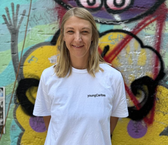 Porträt einer Frau in einem weißen T-Shirt mit youngCaritas-Logo vor einem bunten Graffiti-Hintergrund.