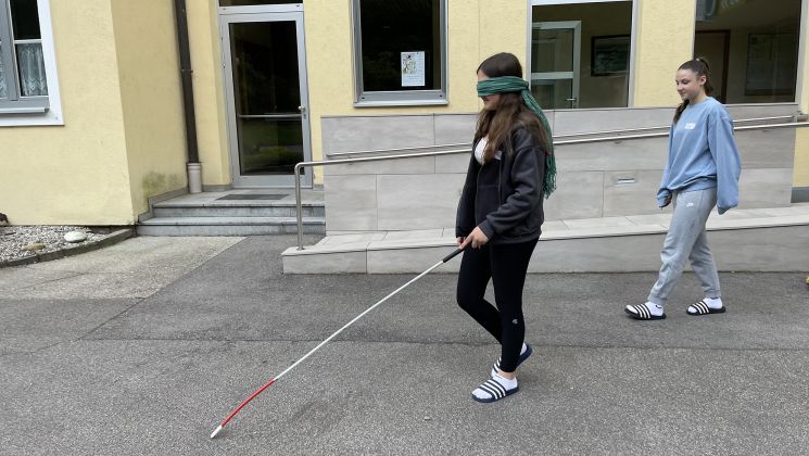 Eine Person mit verbundenen Augen benutzt einen Langstock, während eine andere Person sie begleitet. Sie befinden sich in einem Schulhof.