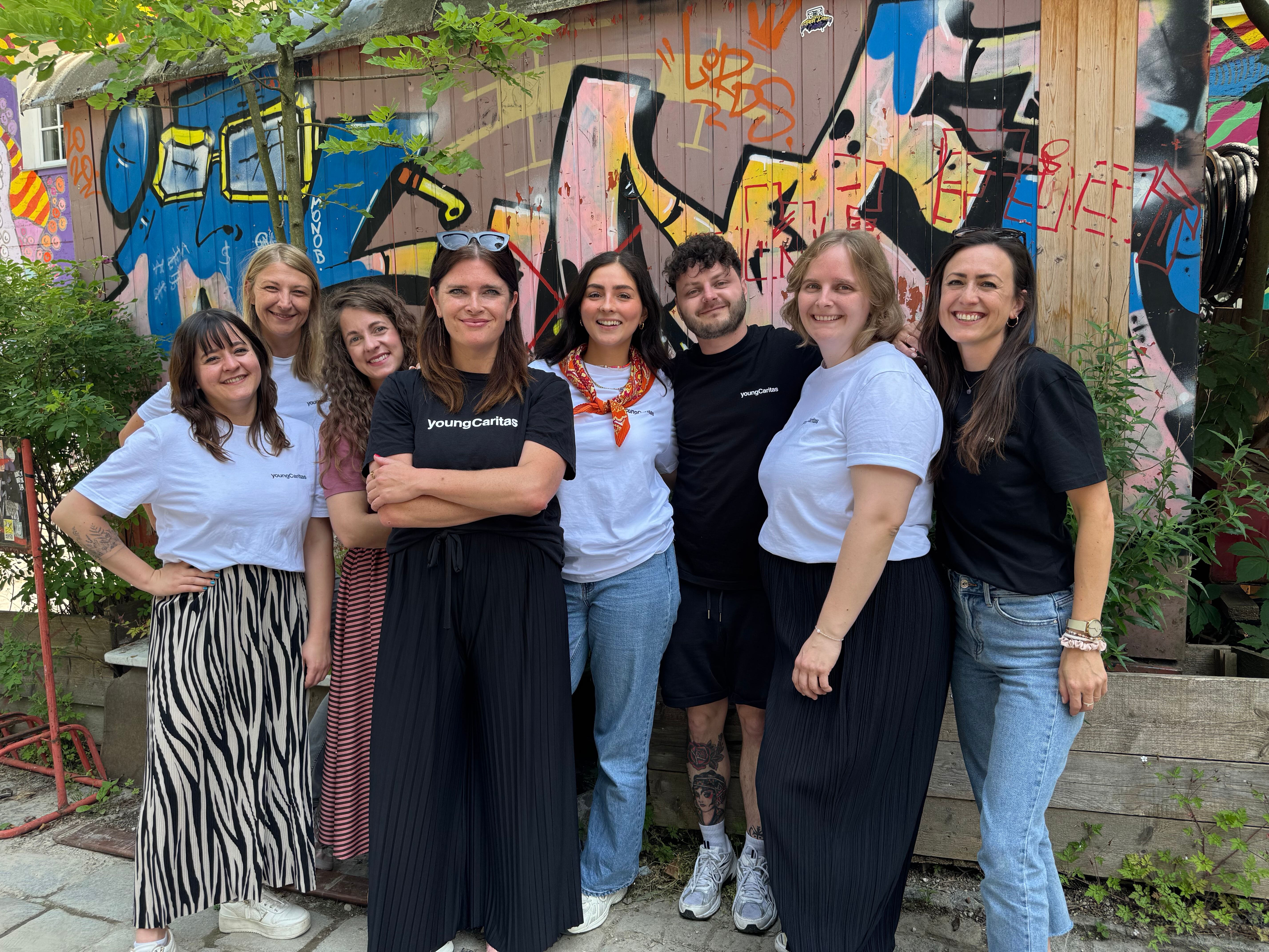 Eine Gruppe von Freiwilligen posiert vor einem bunten Graffiti-Hintergrund. Sie tragen T-Shirts mit dem Logo von youngCaritas.