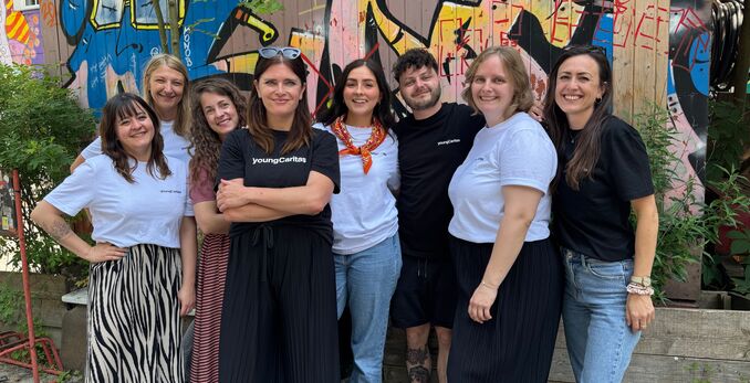 Eine Gruppe von Freiwilligen posiert vor einem bunten Graffiti-Hintergrund. Sie tragen T-Shirts mit dem Logo von youngCaritas.