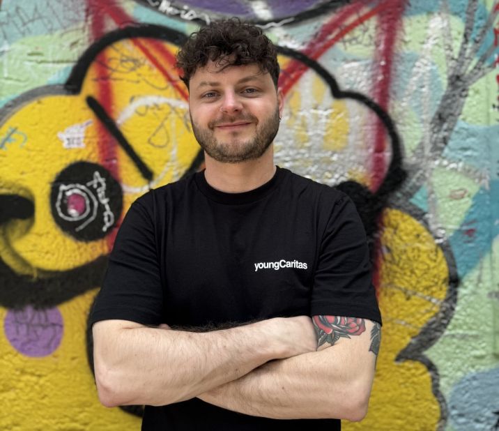 Ein junger Mann mit kurzem, lockigem Haar und einem schwarzen T-Shirt steht vor einer bunten Graffiti-Wand. Auf seinem T-Shirt steht 'youngCaritas'.