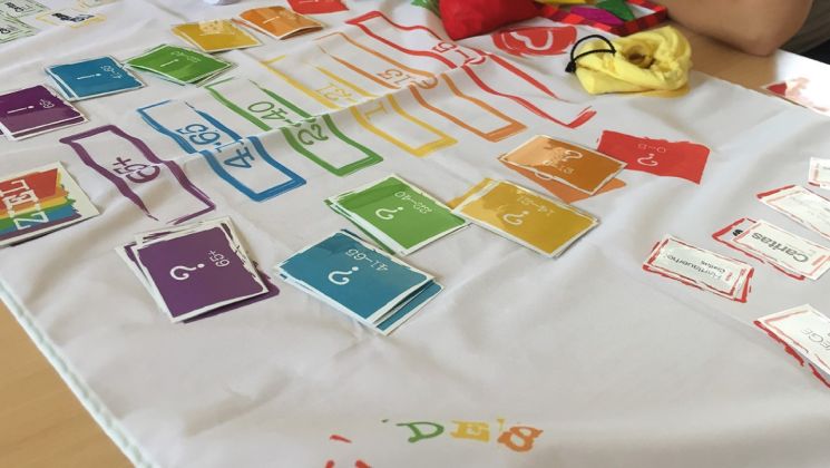 Auf einem Tisch liegt ein Brettspiel mit bunten Karten in verschiedenen Farben und Fragezeichen. Einige Spielsteine und Beutel sind ebenfalls sichtbar.