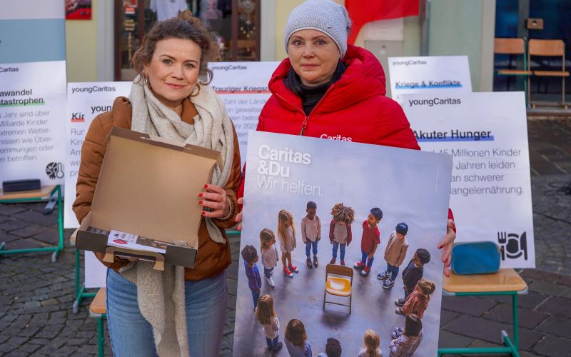 Zwei Frauen präsentieren ein Plakat von Caritas, um auf den Hunger von Kindern aufmerksam zu machen. Hintergrund mit weiteren Ankündigungen.