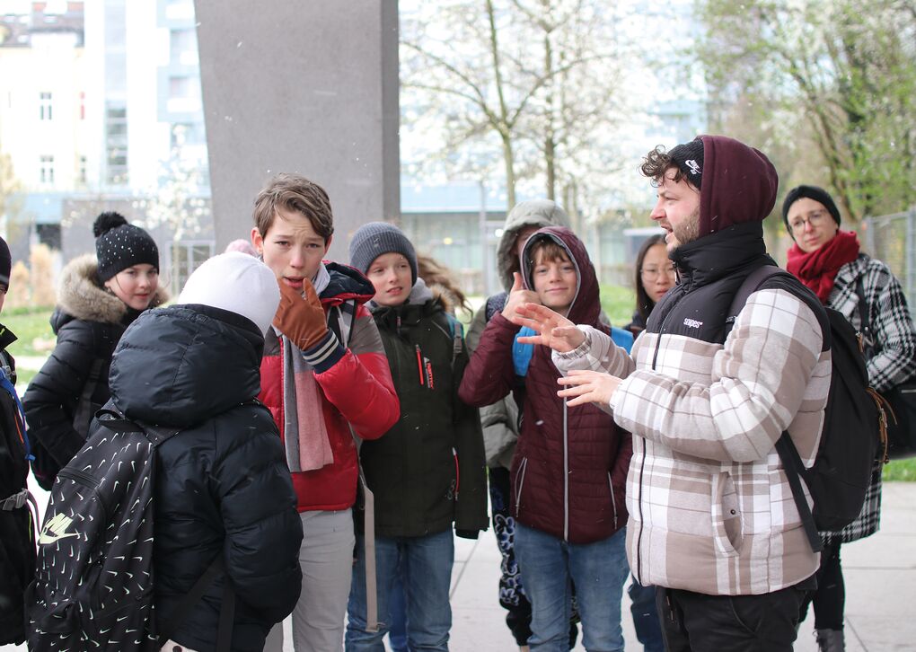 Eine Gruppe von Schülern steht draußen und hört einem Lehrer bei einer Erklärung zu, während sie sich unterhalten.