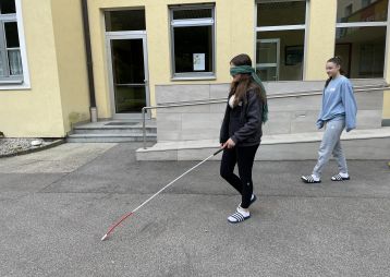 Eine Person mit verbundenen Augen benutzt einen Langstock, während eine andere Person sie begleitet. Sie befinden sich in einem Schulhof.