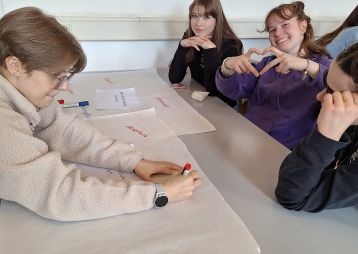 Vier Schülerinnen arbeiten gemeinsam an einem Tisch mit Plakaten und Stiften, während eine von ihnen lächelt und ein Herz formt.