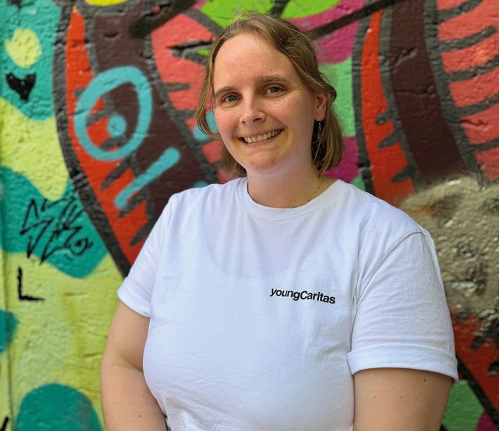 Eine lächelnde Frau in einem weißen T-Shirt mit dem Logo 'youngCaritas' vor einer bunten Graffiti-Wand.