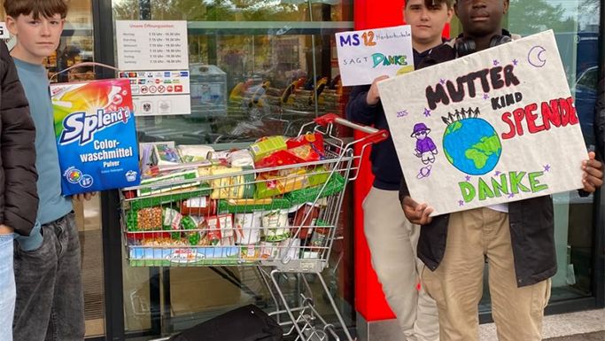 Drei Jugendliche stehen vor einem Supermarkt mit einem Einkaufswagen voller gespendeter Lebensmittel und halten Schilder, auf denen 'Danke' steht.