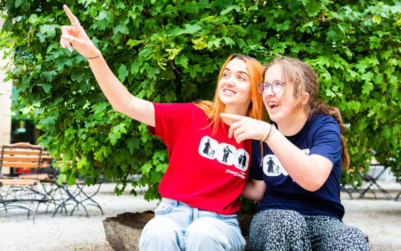 Zwei junge Frauen in lustigen T-Shirts sitzen auf einem Baumstamm und zeigen auf etwas Spannendes in der Umgebung.